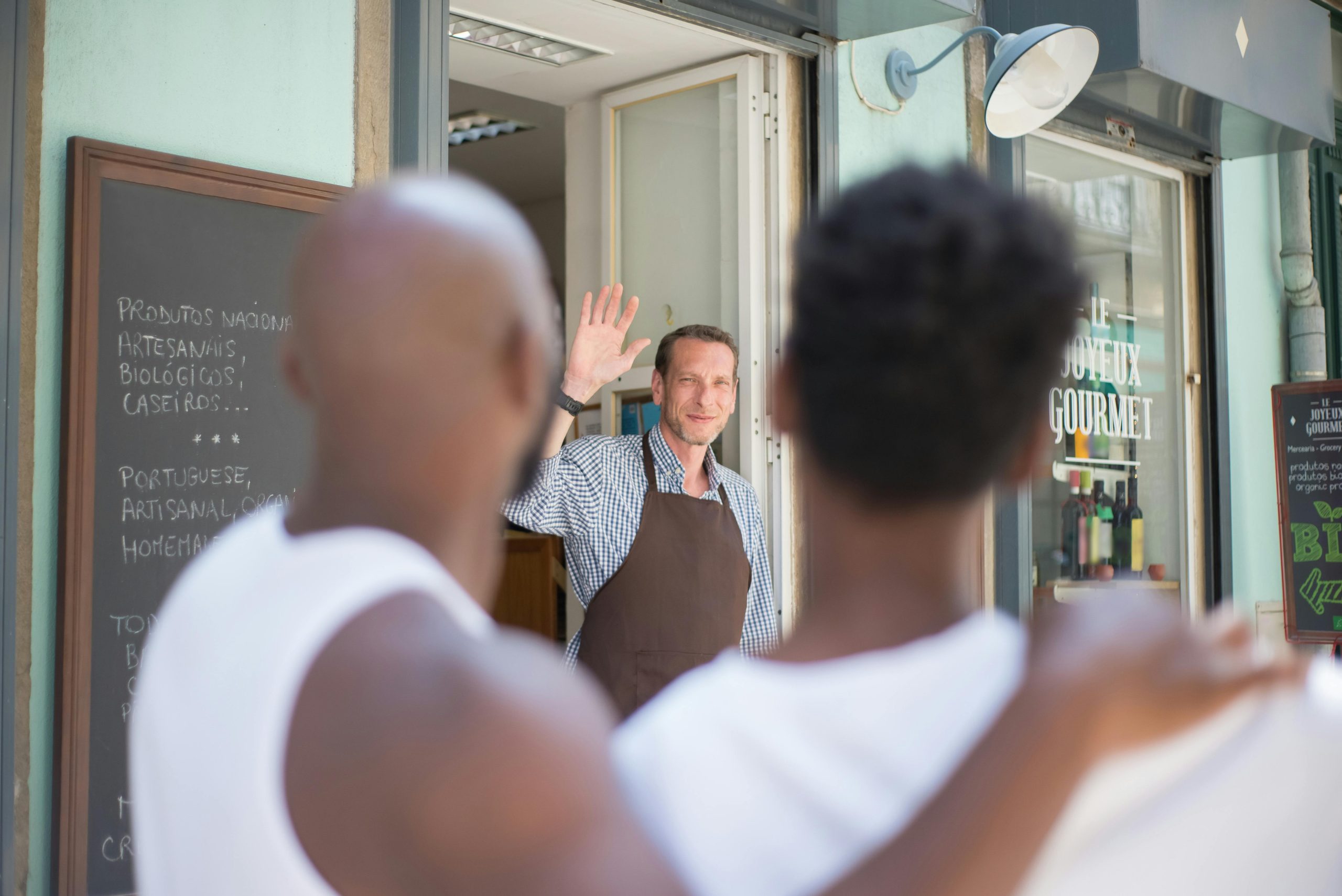 A shop owner standing at the entrance of his store, warmly greeting two customers outside, illustrating community engagement and strong customer relationships.