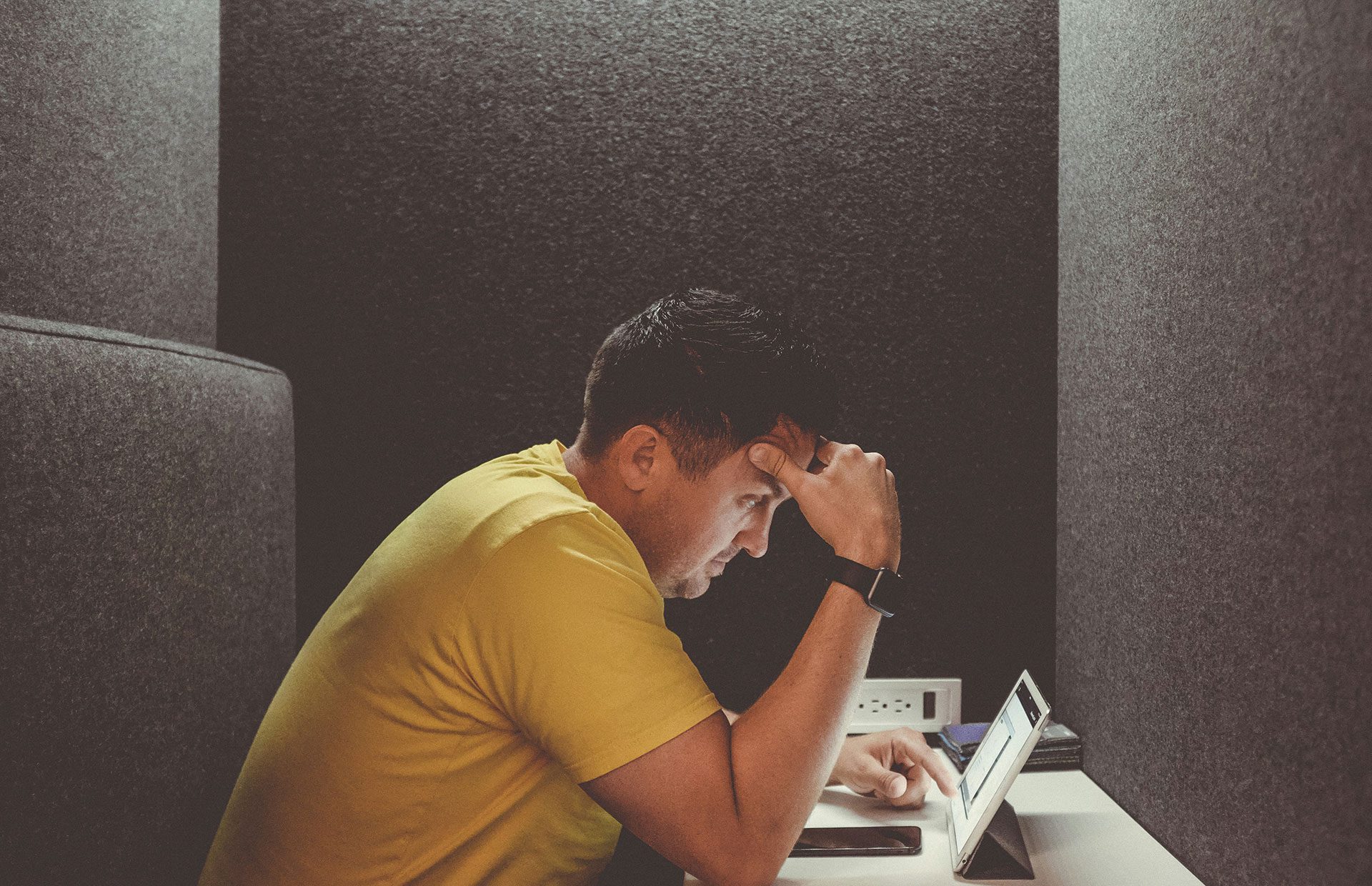 Entrepreneur sitting alone in a confined workspace looking stressed while working on a tablet, representing the pressure and burnout caused by running a business that relies entirely on the founder.